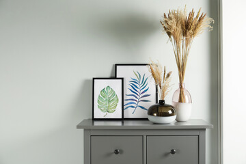 Reed's blossom in glass vases and pictures on grey cabinet indoors. Space for text