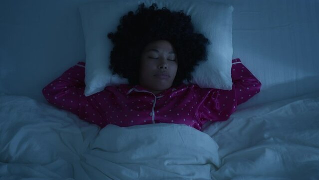 Slow motion face with closed eyes yawning. Beautiful young african american woman with funny afro hairstyle sleeping cozily in comfortable bed on white linens and hugging pillow bedroom at night 4K