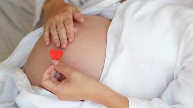 Unrecognizable pregnant woman wearing robe at home caressing,stroking belly, baby bump, holding red heart.Cropped, close-up shot.Happy pregnancy.Love and St Valentine day concept,relaxing having rest.