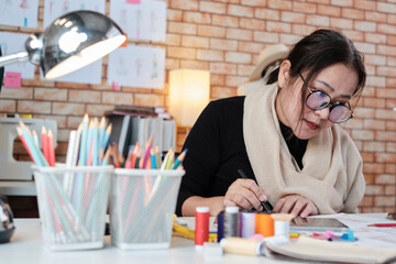 Asian middle-aged female fashion designer works in studio by idea drawing sketches with digital tablet and colorful fabric for a dress design collection, professional boutique tailor SME entrepreneur.