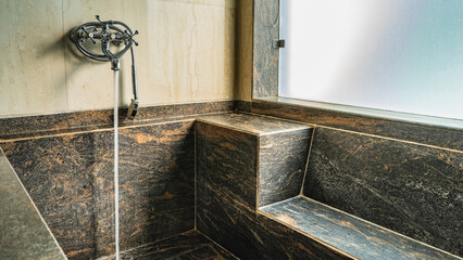 The large bathtub is lined with black marble slabs. There is a seat. A jet of water pours from a...