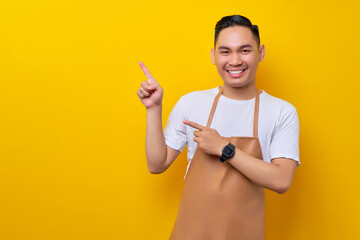 smiling young Asian man barista barman employee wearing brown apron working in coffee shop, pointing finger aside isolated on yellow background. Small business startup concept