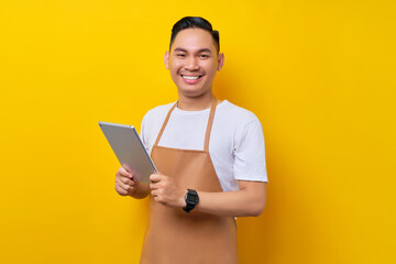 smiling young Asian man barista barman employee wearing brown apron work in coffee shop holding digital tablet and looking at camera isolated on yellow background. Small business startup concept