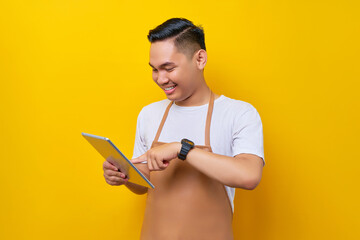 smiling young Asian man barista barman employee wearing brown apron work in coffee shop using digital tablet, browsing the internet on modern gadget on yellow background. Small business startup