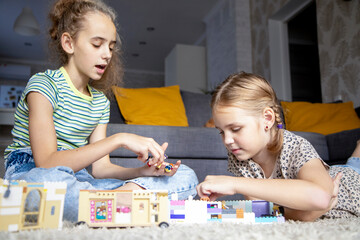 Fototapeta premium Two little sister girls play with multi-colored toy blocks at home in the living room. Educational games for children. Mess in the game room.