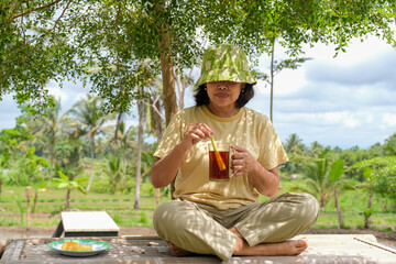 A village girl sitting on bamboo bench in the rice field enjoying some traditional snack and hot...