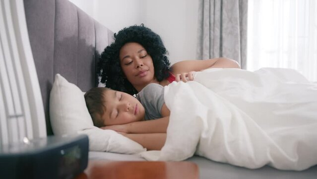 Loving tender African American young mum cover with blanket adorable cute boy, embracing kissing small kid. Happy affectionate mother cuddling with sleeping preschool child son in bedroom at home 4K