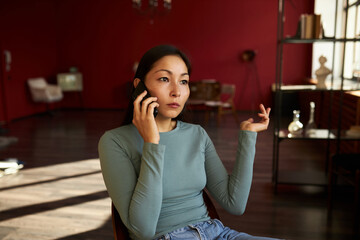Indoor portrait of puzzled astonished asian female talking on phone with her angry husband sitting on chair in her living-room, gesticulating, looking aside with thoughtful face. Unpleasant news