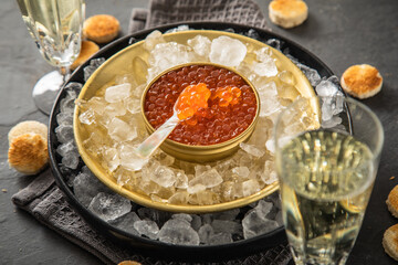 Orange Keta Lachs Kaviar in Dose auf Eis in Glas Schale mit Perlmutt Löffel, Sekt Champagner und Toast Brot auf dunkel Serviette und schwarz Schiefer