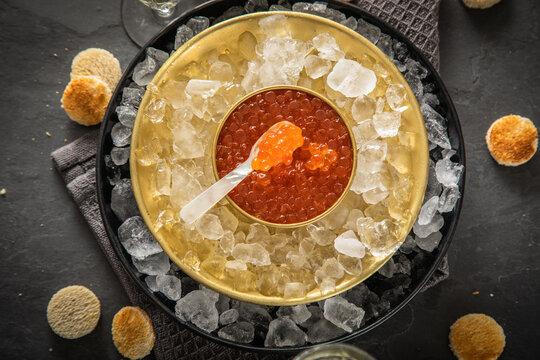 Orange Keta Lachs Kaviar in Dose auf Eis in Glas Schale mit Perlmutt Löffel, Sekt Champagner und Toast Brot auf dunkel Serviette und schwarz Schiefer