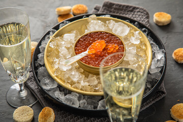 Orange Keta Lachs Kaviar in Dose auf Eis in Glas Schale mit Perlmutt Löffel, Sekt Champagner und Toast Brot auf dunkel Serviette und schwarz Schiefer