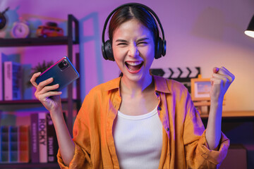 Excited Young Asian woman playing an online game on a smartphone with fists clenched celebrating victory expressing success. - obrazy, fototapety, plakaty