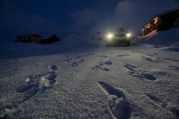 Fotobehang Street of residential area of Selfoss completely snow covered, with human footprints and a 4x4 jeep parked next to a house and with lights illuminating the tracks. © sirbouman
