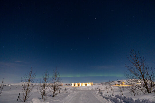 Northern lights on the horizon line and stars with a state of the art illuminated house at the end of a totally snowy road illuminated by moonlight in Iceland with tree silhouettes.