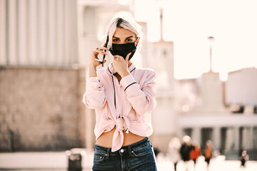 Fototapeta premium Woman with face mask looking at camera while talking on the phone outdoors.