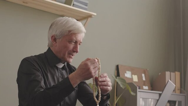 Caucasian senior priest with rosary beads in hands having video conversation with parishioner via laptop, working from home office