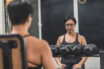 Fototapeta na wymiar A fit middle aged asian woman with short hair getting into the zone before a workout. Resting in between sets. Looking at the mirror at the gym.