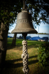 Fototapeta premium Ships bell removed from its vessel now hangs in a garden. The bell has no name only a date 1827. it is now on land but close to water, sail boats can be seen in the background floating on a lake