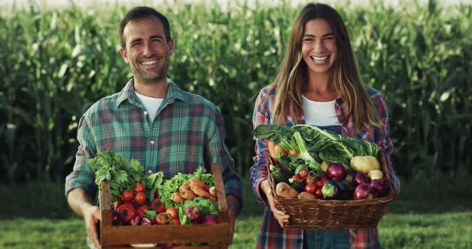  Portrait of a Cheerful Young Couple Holding a Box Full of Vegetables, Looking at the Camera with a Greenery Background and Smiling. Female and Male Farmers Happy with their Harvest of the Year