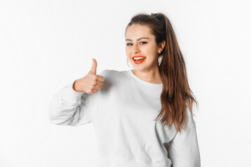 Excited smiling young brunette woman with long hair, wear white sweatshirt show thumb up. Smiling girl looks cheerful and joyful, stands against white studio background