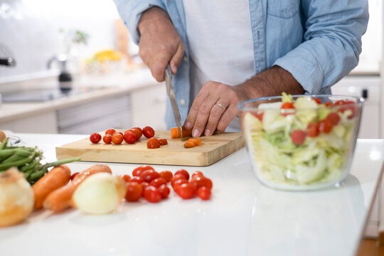 Close up of man at home preparing vegetables salad for lunch. People in the kitchen cooking and enjoying healthy food lifestyle. One unrecognizable male cutting tomatoes cherry to eat alone