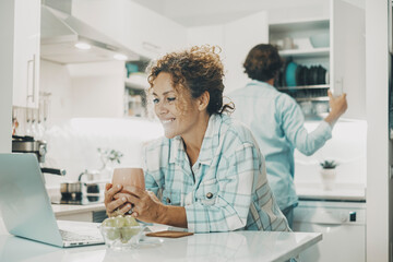 Obraz premium Young adult couple at home in the kitchen busy in indoor leisure domestic activity together. Woman watching a laptop for fun and man preparing lunch in background. Happy life and relationship