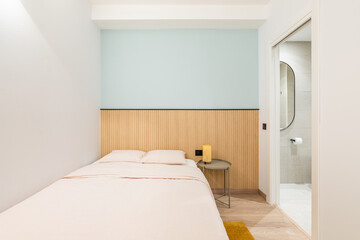 Double bed against a shallow niche with a blue wall and wooden decor. In the bedroom, there is an open door overlooking a bright toilet room with a toilet bowl and a small plastic window.