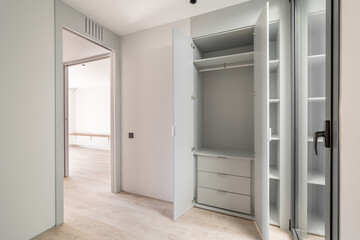 Spacious entrance hall in light cream shades. An elegant front glass door in a black metal frame adds zest to the interior. Open spacious white wardrobe with chest of drawers inside.