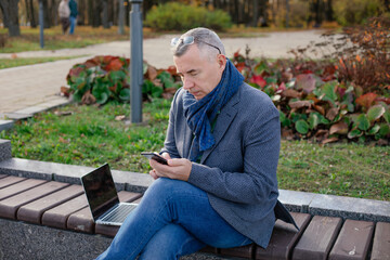 Aged gray haired businessman with glasses sit on bench in autumn city park working on laptop and chatting on telephone.