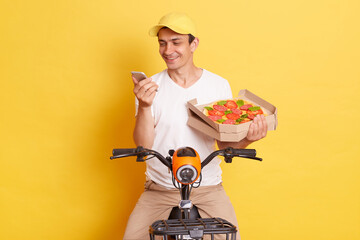Smiling positive delivery man with fresh baked pizza in carton box, wearing white t-shirt, sitting on fast motorbike and using mobile phone while waiting client isolated over yellow background.