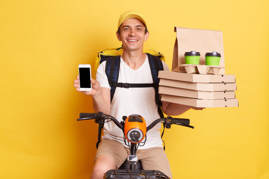 Image of handsome delivery man showing pizza in cardboard box, coffee to go, and smart phone with empty area for advertisement, sitting on fast motorbike isolated over yellow background.