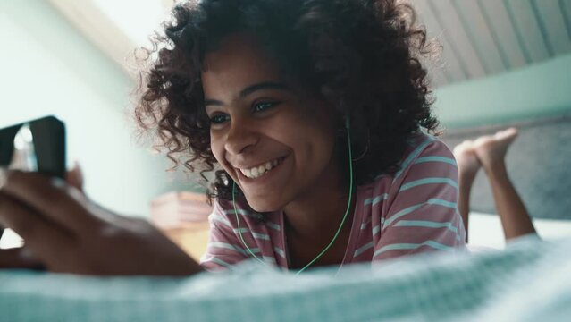 Cheerful curly haired young African girl talking by video call on mobile in headphones on the bed at home