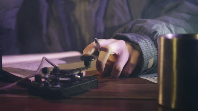 Man transmits information in Morse code by telegraph sitting at table. Person sends signals by tapping finger and holding pen in hand closeup