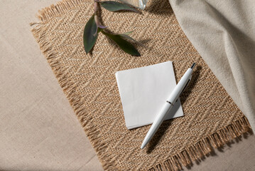 Blank paper, invitation card sheet with empty mock up copy space, pen and plant on neutral beige jute placemat and linen cloth