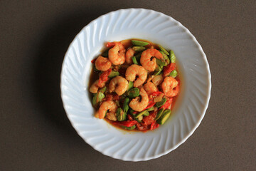 Chili prawns or Sambal Udang with petai or pete or mlanding (Parkia speciosa) cooked with mashed red chili and tomato in white plate, on black background. Indonesia cuisine food, top view.
