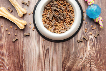 A bowl with dog food, dog treats and toys on a wooden floor.