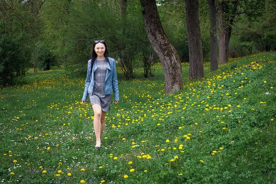 A positive, beautiful girl in a gray dress and a blue jacket, with glasses on her hair, walks through a meadow with grass and small white and yellow flowers during the day.