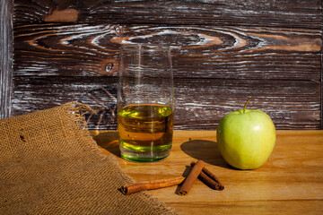 A glass of freshly squeezed apple juice, cinnamon sticks and a green ripe apple on a table covered...