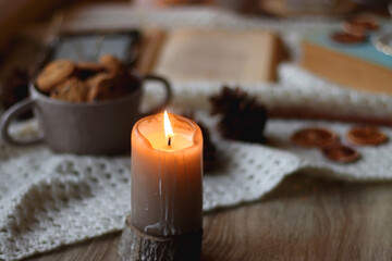 Cup of tea, books, tablet, bowl of cookies, various spices, pine cones and lit candles. Hygge at home. Selective focus.