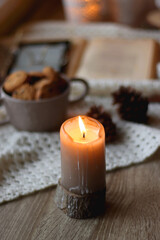 Cup of tea, books, tablet, bowl of cookies, various spices, pine cones and lit candles. Hygge at home. Selective focus.