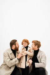 happy redhead kid in trench coat smiling near father and bearded grandpa hugging him isolated on grey.