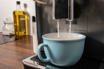 Making coffee. Milk flows from the coffee machine into a blue cup, kitchen table in the background