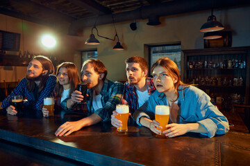 Group of young people, men and women, sport fans watching match at pub. Attentively looking, emotionally cheering up favourite team.