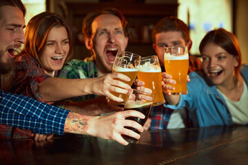 Group of young people, men and women watching match at pub, drinking beer. Fans emotionally cheering up favourite sport team. Happy