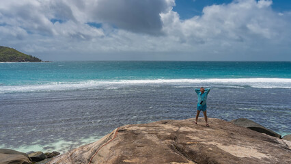 A man stands on the edge of a granite cliff, above the ocean, smiling. His arms are thrown behind his head, clothes are fluttering in the wind. Foam of waves on turquoise water. Clouds in the blue sky