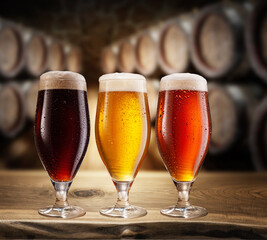 Three type of beer with foam head on wooden table. Blurred brewery cellar at the background.