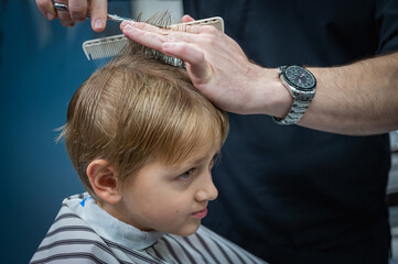 Fototapeta premium Happy cute fair-haired preschool boy getting a haircut. Children's hairdresser with scissors and comb cuts a little boy's hair in a room with a loft interior