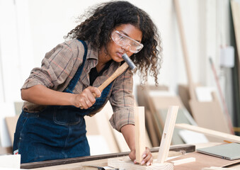 Young female carpenter is working in woodwork DIY furniture in carpentry workshop. Multiracial woman woodworker busy using hammer to assemble DIY furniture at workbench. Joiner and craftsman concept.