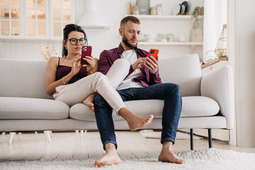Young caucasian couple in casual sits on couch at home using phones, searching new apartment via internet. Relocation. Serious man at social media. Changing real life for virtual. Marriage troubles. - obrazy, fototapety, plakaty