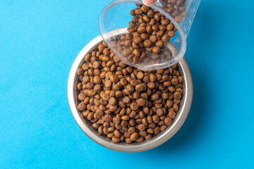 Pouring ration dry pet food in a metal bowl on a blue background. Feeding cats and dogs with granulated dry food. Pellets of healthy food for dogs and cats. Healthy food for pets